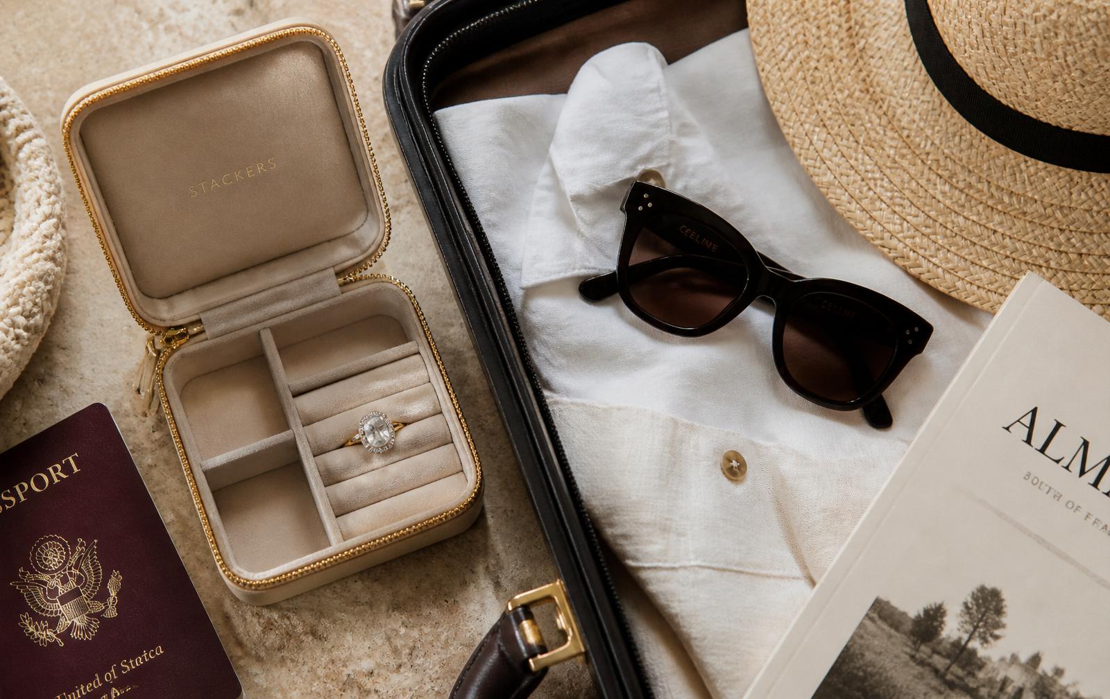 A packed suitcase with white linen, sunglasses and an open jewellery travel case